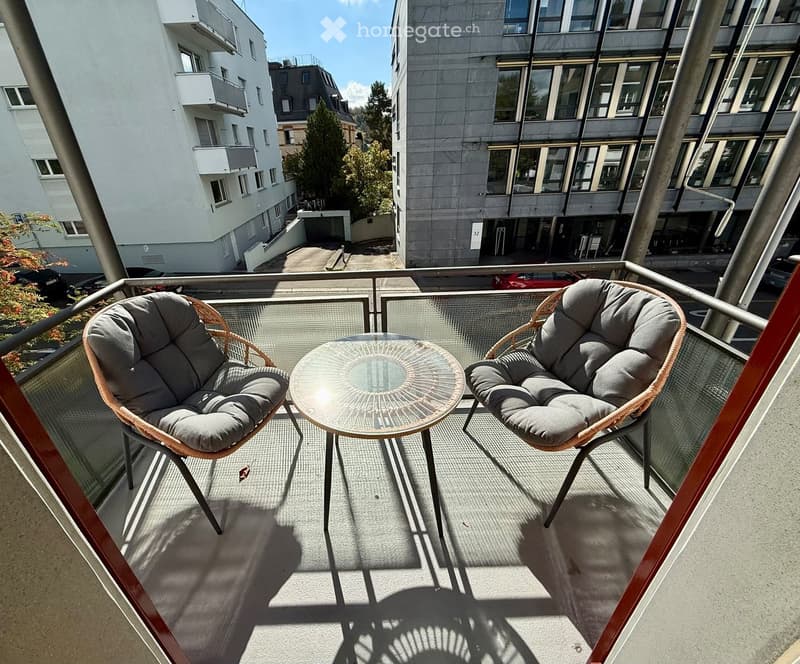 Balcony with two wicker chairs and a round glass table, overlooking a modern residential area with buildings and trees.
