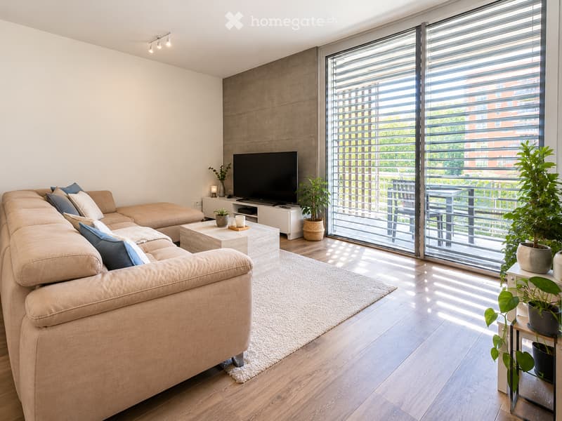 Modern living room with a large sectional sofa, coffee table, TV, side table, and potted plants. Large windows with blinds lead to a balcony.