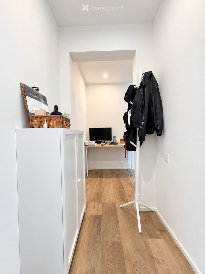 A narrow hallway with wooden flooring, a small desk with a computer, a coat rack with jackets, and a wicker basket on top of a cabinet.