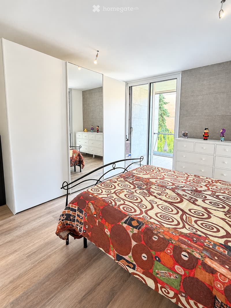 Modern bedroom with wooden flooring, a bed with a colorful patterned bedspread, a wardrobe, a dresser, and a mirror. The room features large windows and a sliding glass door leading to a balcony.