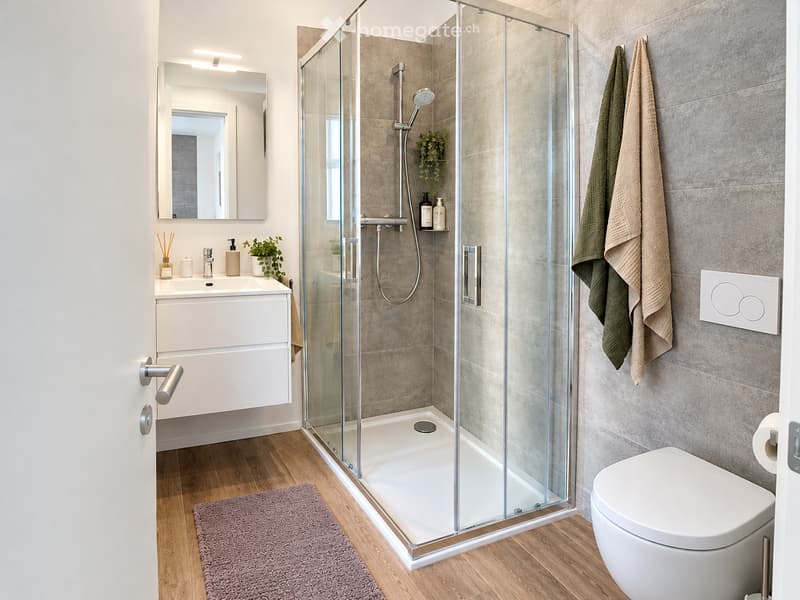 Modern bathroom with a glass-enclosed shower, white sink with mirror, toilet, and towels hanging on the wall. The space features wooden flooring and a sleek, contemporary design.