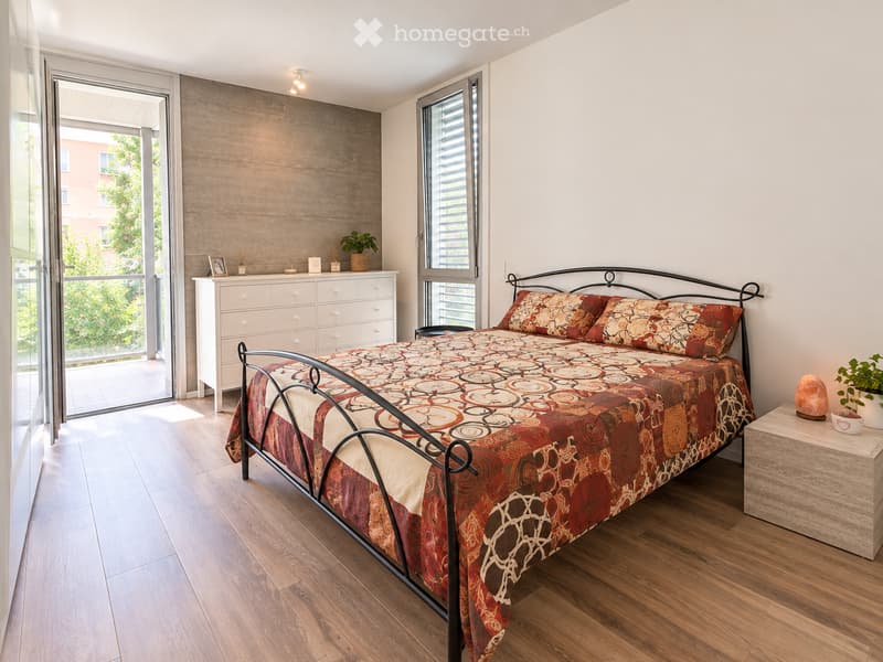 Modern bedroom with wooden flooring, large windows, and a balcony. Features a metal-framed bed with patterned bedding, a white dresser, and a small nightstand with a plant.