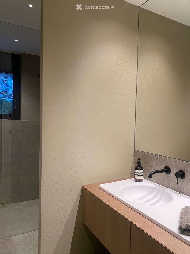 Modern bathroom with a sleek white sink, black faucet, soap dispenser, and a folded towel on the counter. The walls are light-colored with a textured finish, and the floor is tiled.