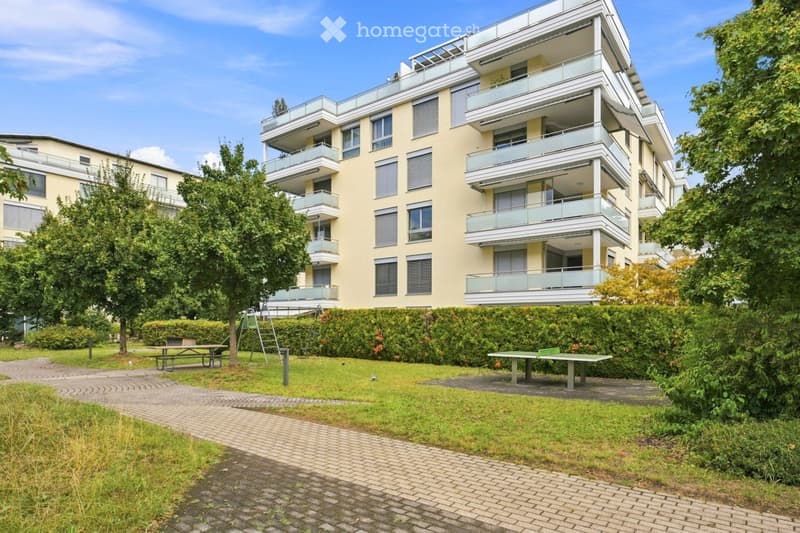 Modern multi-story residential building with balconies, surrounded by greenery and recreational area with picnic tables and a swing set.