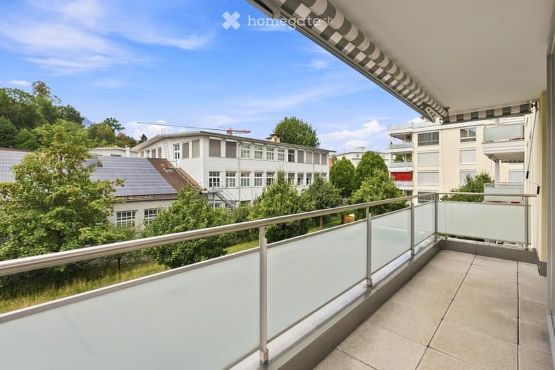 Balcony with glass railing, tiled floor, and striped awning, overlooking a green area with trees and buildings.