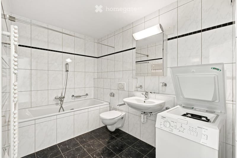 Modern bathroom with white tiles, bathtub, toilet, sink, mirror, and a washing machine. Features a glass shower enclosure and a towel rack.