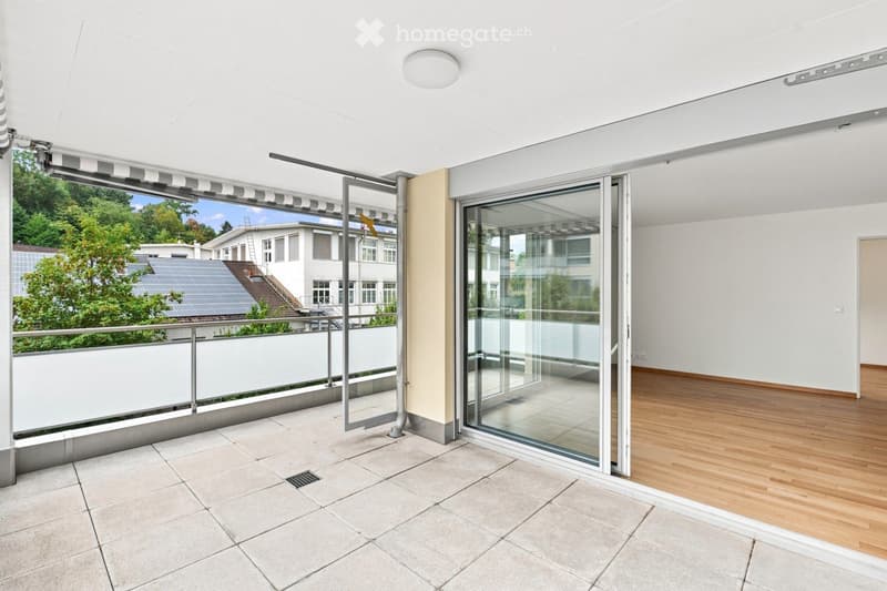 Modern balcony with large glass sliding doors, tiled floor, white railing, and a retractable awning. The view shows a green area with trees and neighboring buildings.