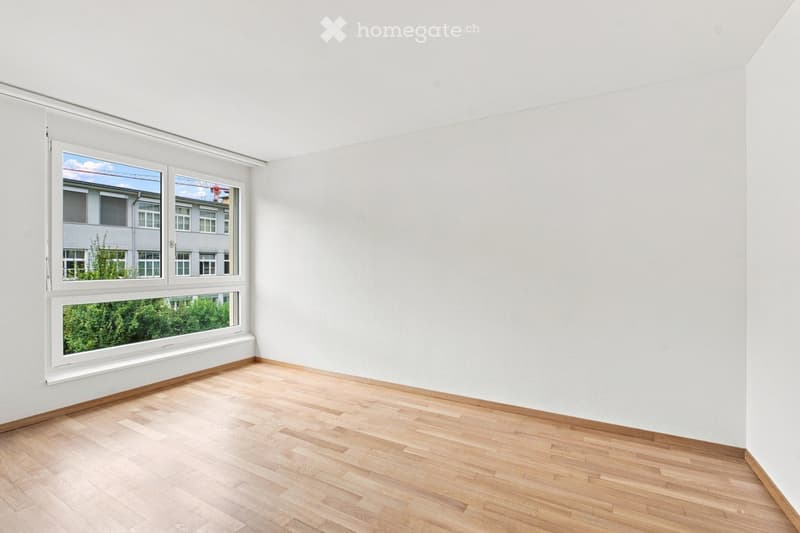 Empty room with light wooden flooring and white walls, featuring large windows with white frames that offer views of buildings and greenery outside.