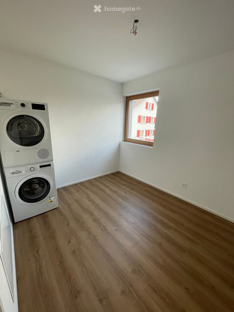 A small room with light wooden flooring and white walls, featuring a stacked washing machine and dryer unit against one wall and a window showing a view of a neighboring building.