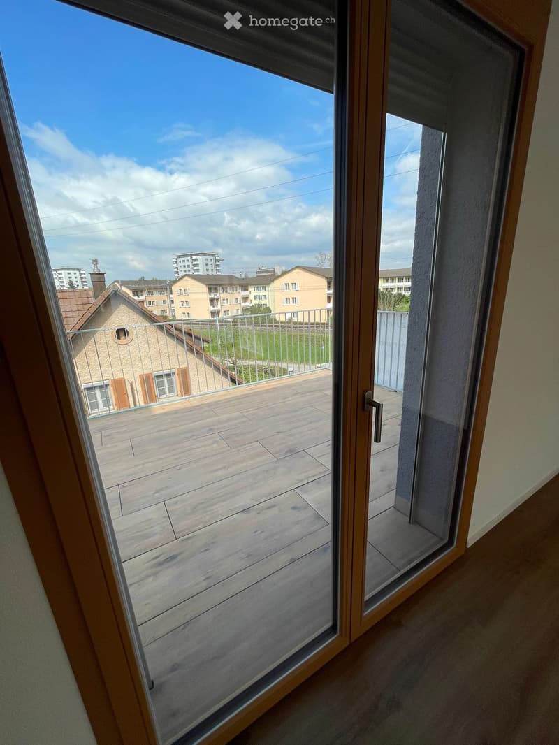 Balcony with wooden decking, metal railing, and view of surrounding buildings and greenery.