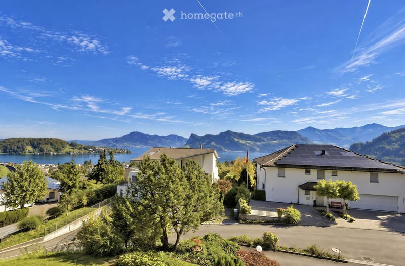 Two-story house with solar panels on the roof, surrounded by trees and a scenic lake and mountain view in the background.