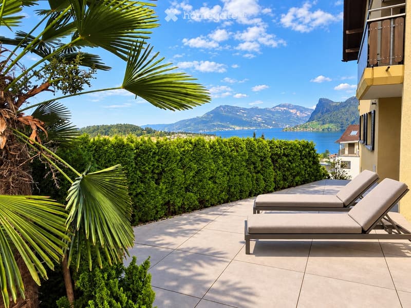 Balcony with sun loungers, surrounded by lush greenery and offering a scenic view of a lake and mountains.