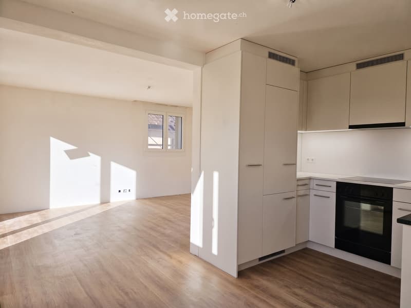 Empty kitchen with white cabinets, wooden flooring, and a window letting in natural light. No appliances or furniture are present.