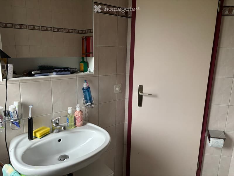 A small bathroom with a white sink, faucet, toothbrush, toothpaste, soap dispenser, and a mirror above the sink. Shelves hold various toiletries and a towel is visible on the floor.