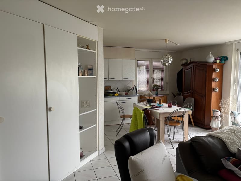 A modern, open-concept living and dining area with a kitchen in the background. The living area features a black leather sofa with white and red pillows, a wooden coffee table, and a dining table with chairs. The kitchen has white cabinets, a countertop w