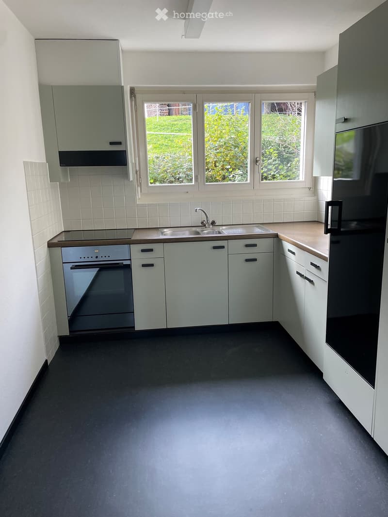 Modern kitchen with light grey cabinets, stainless steel sink, oven, and refrigerator. The space features a dark grey floor and white tiled backsplash.