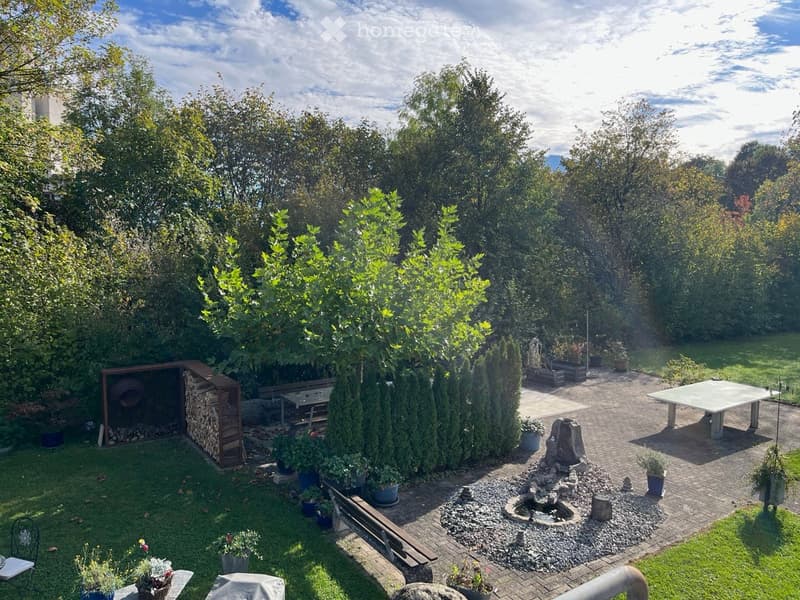 Well-maintained garden with a mix of lawn, paved pathways, and various plants. Features include a wooden bench, a small fountain, a table, and a storage shed for firewood.