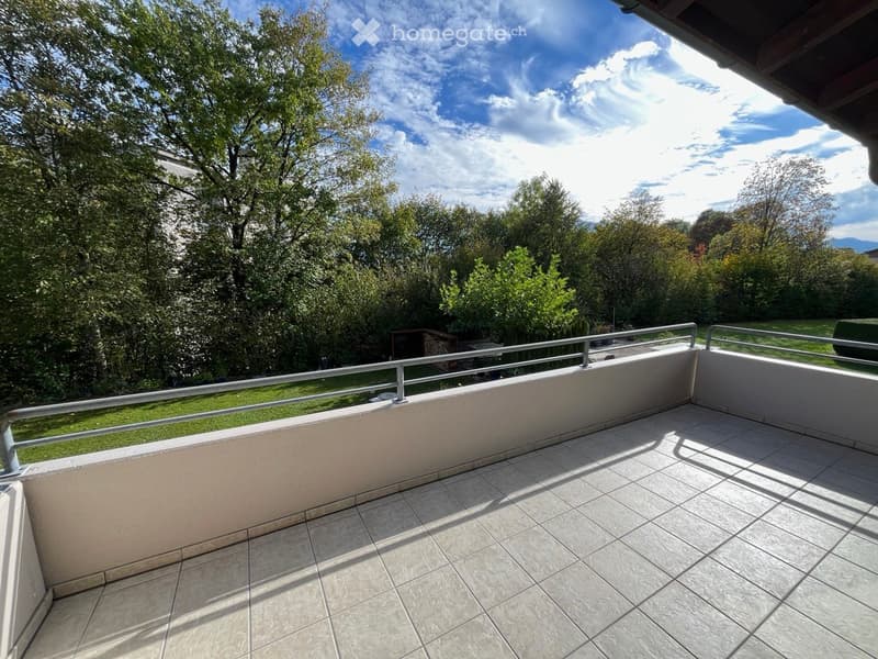 Balcony with tiled floor, metal railing, and a view of a green, tree-filled area under a partly cloudy sky.