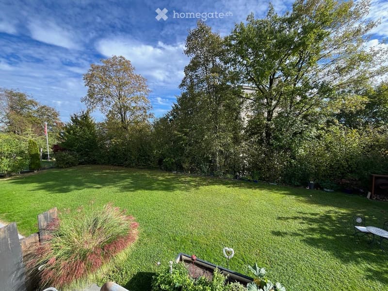 Well-maintained garden with a large lawn, trees, shrubs, and a small seating area with a table and chairs.