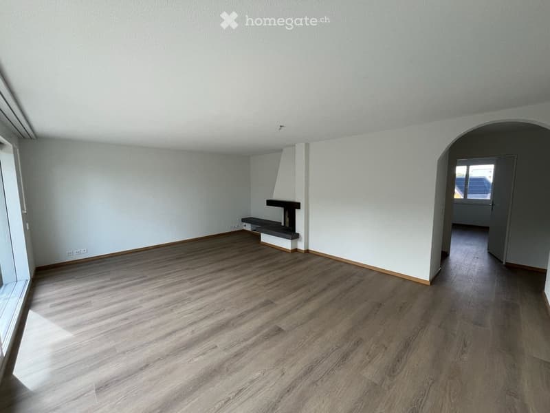 Empty room with light-colored wooden flooring, white walls, and a small built-in bench and fireplace. The room has large windows allowing natural light to enter.