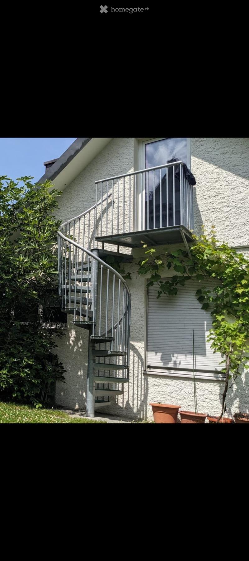 Spiral staircase leading to a balcony with a metal railing, surrounded by greenery and potted plants.