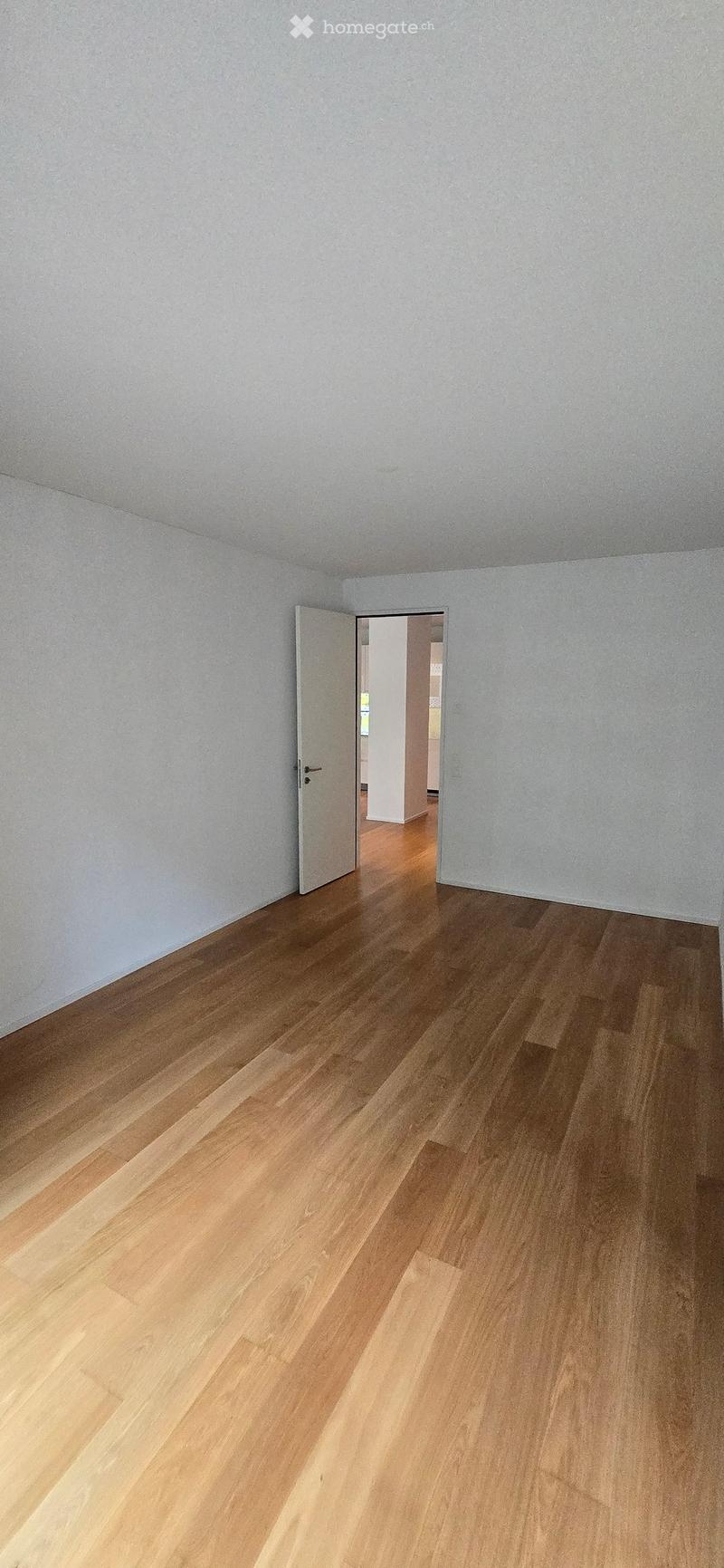 Empty room with light wooden flooring and white walls, featuring an open doorway leading to another room.