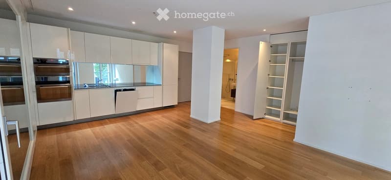 Open-plan unfurnished room with light wooden flooring, white cabinetry, and built-in shelving. The space includes a kitchen area with integrated appliances and a separate storage area with open shelves.