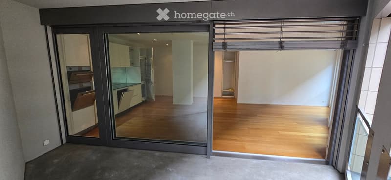 Empty room with wooden flooring, large sliding glass doors leading to another room with kitchen appliances, and a closet area.