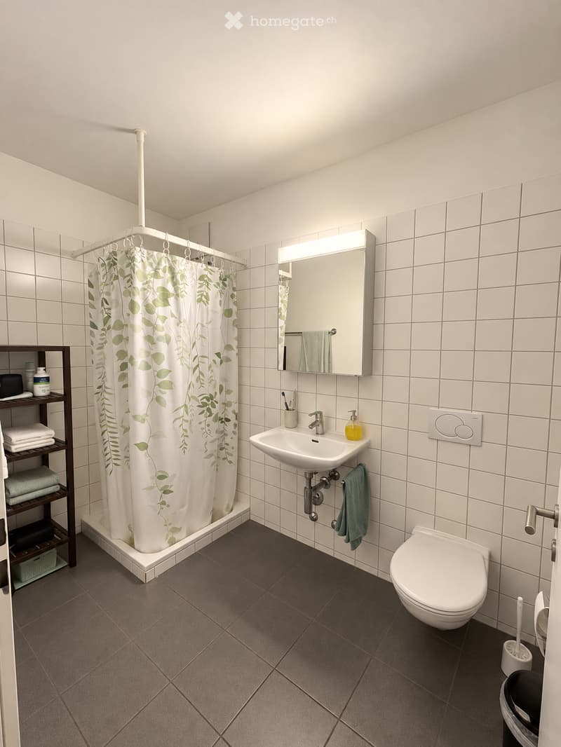 Modern bathroom with white tiled walls, dark tiled floor, a white sink with a mirror, a toilet, a shower with a leaf-patterned curtain, and a small wooden shelf holding towels and toiletries.