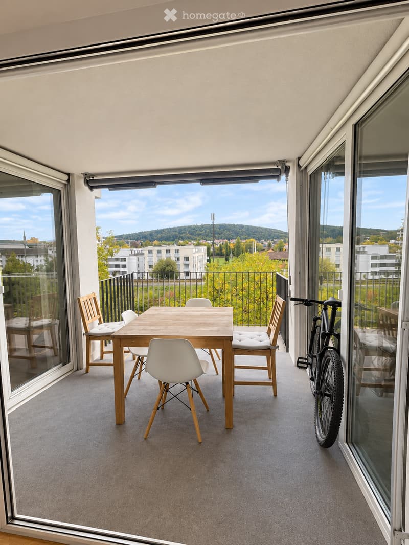Balcony with a wooden table, four chairs, and a bicycle, offering a scenic view of the city and surrounding landscape.