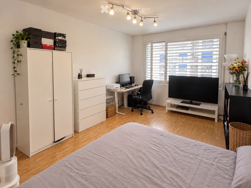 Modern bedroom with a bed, wardrobe, desk, chair, TV, and a flower vase on a side table. The room features wooden flooring and large windows with blinds.