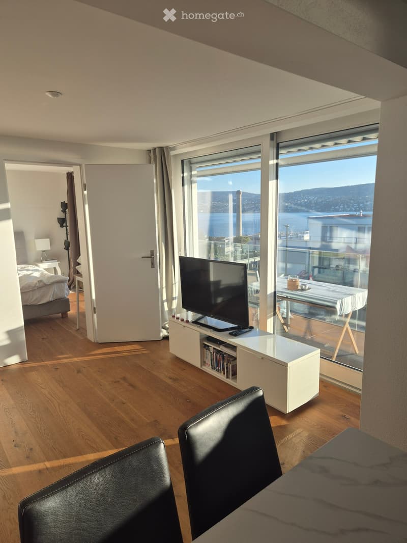 Modern living room with large windows offering a scenic view, featuring a leather chair, a white cabinet with a TV, a bed in the adjacent room, and a nightstand with a lamp.