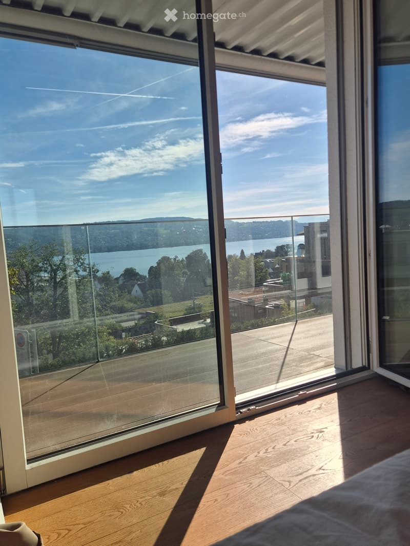 Bright bedroom with wooden flooring, large glass doors opening to a balcony with scenic lake and mountain views.
