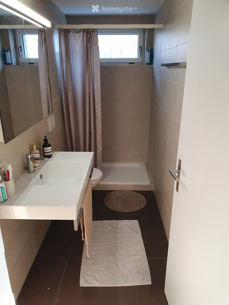 Modern bathroom with a white sink, toilet, bathtub, and mirror. Beige tiles, dark floor, and two towels on the floor.