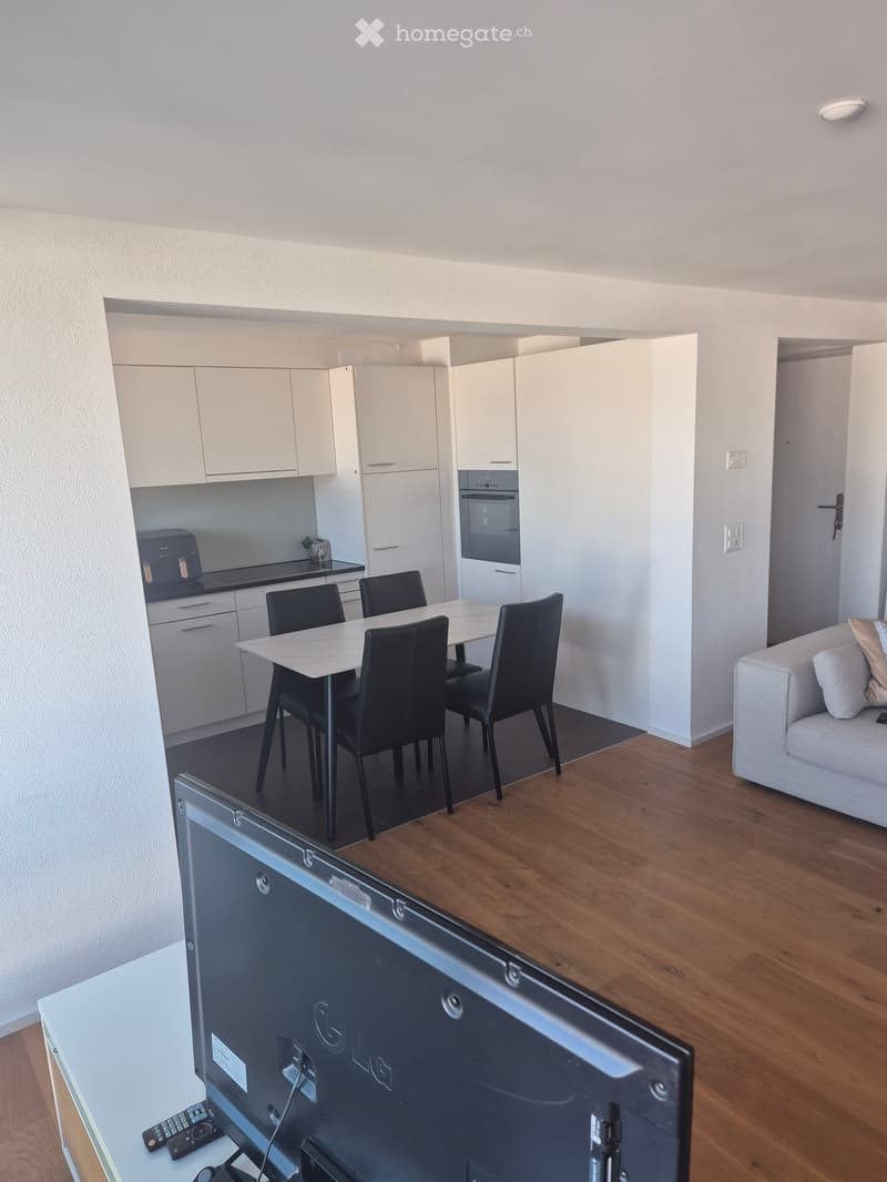 Open-plan living and dining area with a sofa, dining table with chairs, and a television on a stand. The kitchen area features white cabinets and an oven.