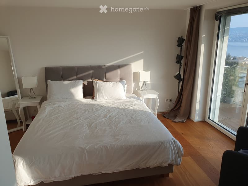 Modern bedroom with a large bed, nightstands, lamps, and a view of the sea through large windows.