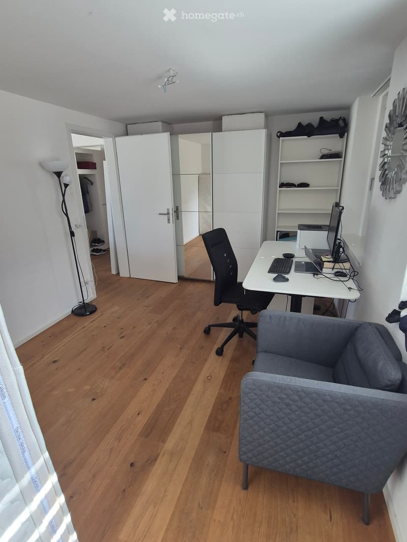 A modern, well-lit room with wooden flooring, a grey sofa, a white desk with a laptop, keyboard, and mouse, a black office chair, a tall floor lamp, and built-in white shelving and wardrobe units.