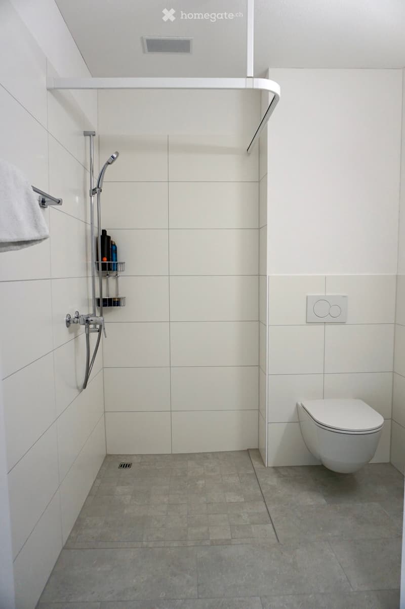 Modern bathroom with white tiled walls, grey tiled floor, toilet, showerhead, towel, and shower rack.