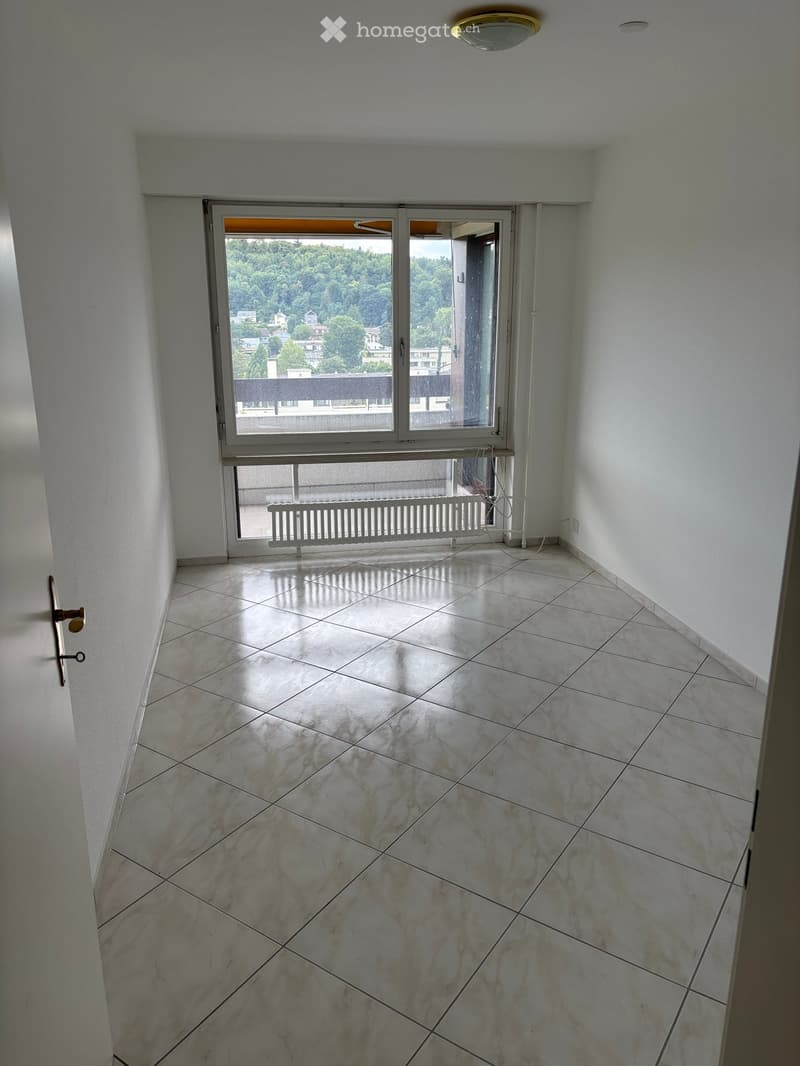 Empty room with tiled floor, white walls, and a large window opening to a balcony with a view of greenery and buildings.