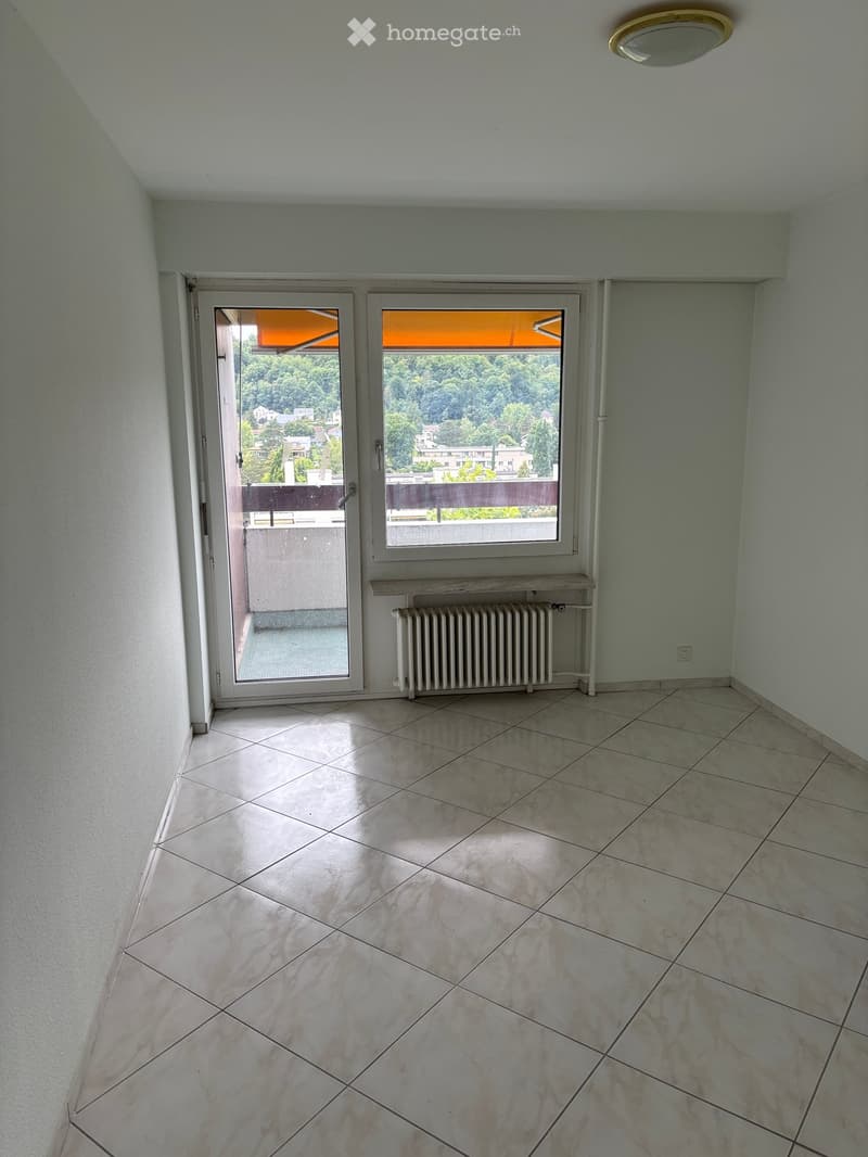 Empty room with tiled floor, light-colored walls, a radiator, a small shelf, and a sliding glass door leading to a balcony with a view of greenery and buildings.
