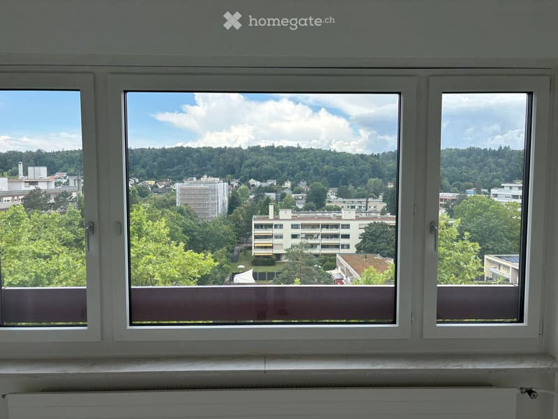 Balcony with large windows offering a view of a cityscape, trees, and hills under a partly cloudy sky.