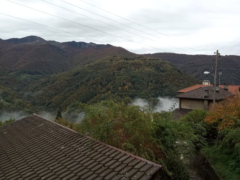 A scenic outdoor view from a rooftop, featuring a house with a terracotta roof, lush greenery, and misty mountains in the background.