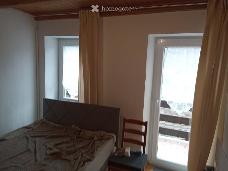 Bedroom with a bed, a chair, and a small table near the window. The room has wooden beams on the ceiling and a sliding glass door leading to a balcony.