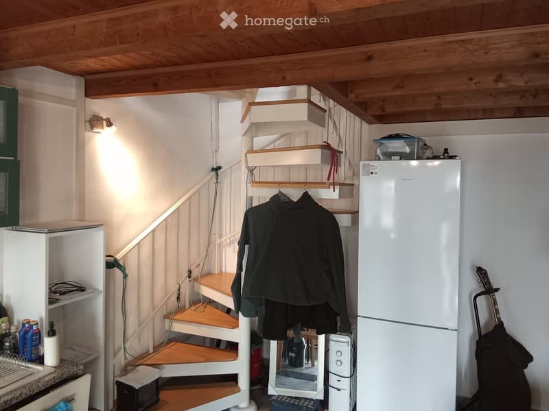 A small, cozy room with a staircase leading upwards, a refrigerator, a laptop on a shelf, a guitar leaning against the wall, and various small items on a counter.