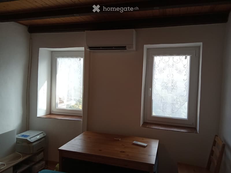 A small, unfurnished room with wooden table, chair, laptop, and printer. Two windows with decorative glass panes and an air conditioning unit mounted on the wall.