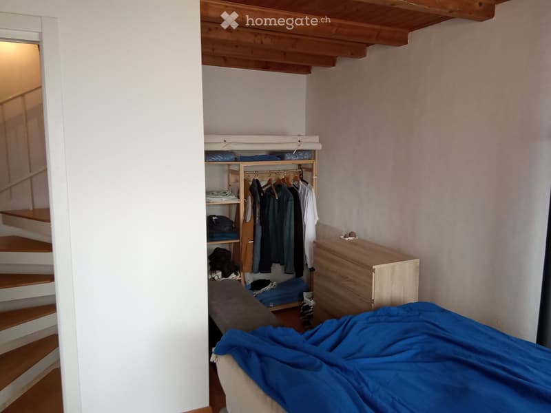 Cozy bedroom with a bed covered in blue bedding, a wooden wardrobe with hanging clothes, a small wooden drawer, and a shelf with folded blankets and other items. Wooden staircase leads to an upper level.
