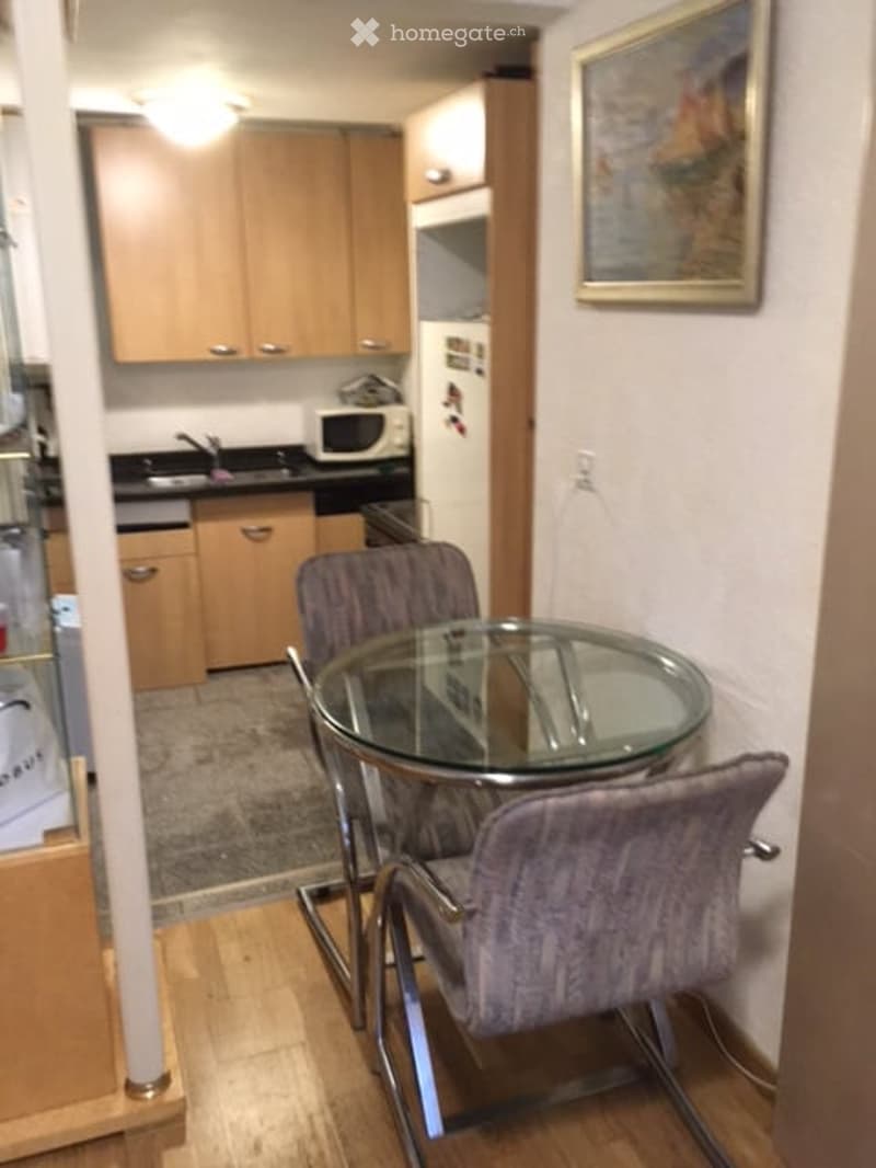 A compact, well-lit kitchen and dining area with light wooden cabinets, a glass dining table with four upholstered chairs, and a microwave on the counter.