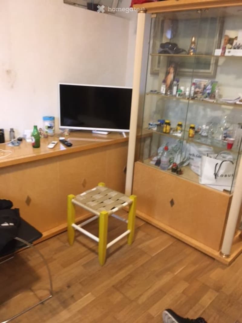 A small, furnished room with a wooden floor, a glass-fronted cabinet, a small stool, a chair, and a screen on a wooden cabinet.