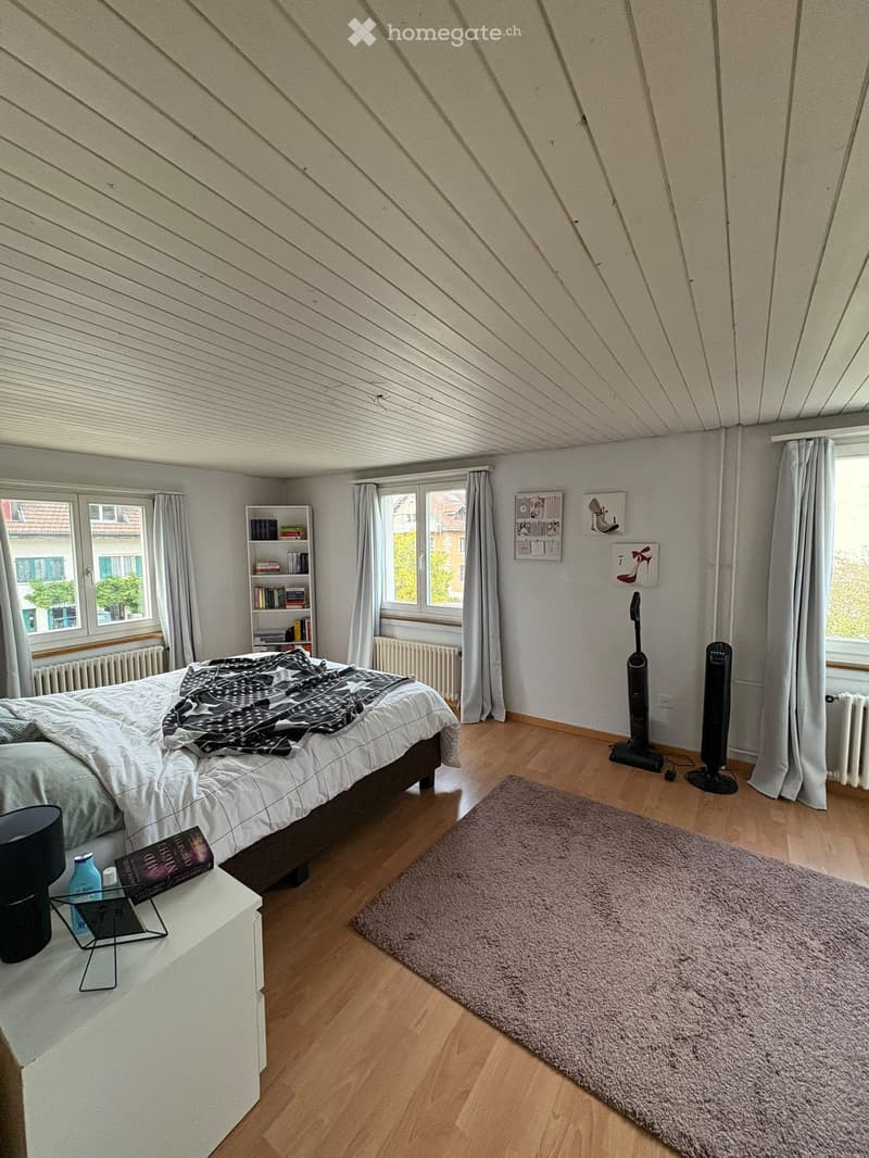 Bright bedroom with wooden flooring, slanted ceiling, large windows with curtains, bed with bedding, nightstand, bookshelf, and decorative items on walls.