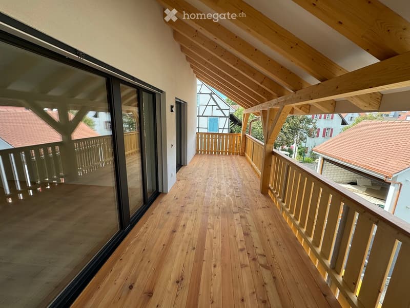 Wooden balcony with slanted roof, large glass window, wooden railing, and view of neighboring houses.